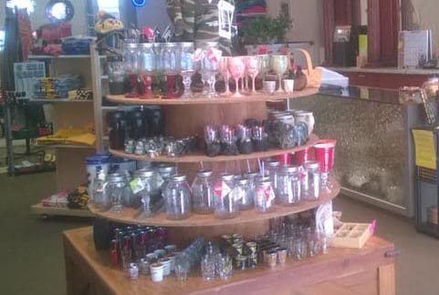 A retail store display featuring a variety of glassware, including jars, glasses, and cups arranged on a multi-tiered wooden table. The shelves around the display hold various other products, and the room is well lit with natural light from windows.