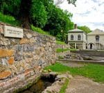 A historic site surrounded by lush greenery features a stone wall with a sign reading "George Washington's Bath Tub (1749)." Behind the wall, there are two old, cream-colored buildings with a metal roof and lush green trees in the background.