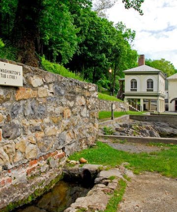 A historic site surrounded by lush greenery features a stone wall with a sign reading "George Washington's Bath Tub (1749)." Behind the wall, there are two old, cream-colored buildings with a metal roof and lush green trees in the background.