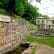 A historic site surrounded by lush greenery features a stone wall with a sign reading "George Washington's Bath Tub (1749)." Behind the wall, there are two old, cream-colored buildings with a metal roof and lush green trees in the background.