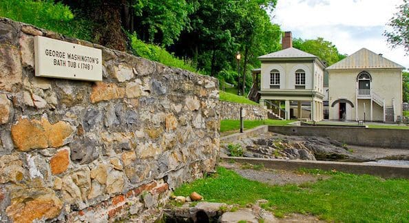 A historic site surrounded by lush greenery features a stone wall with a sign reading "George Washington's Bath Tub (1749)." Behind the wall, there are two old, cream-colored buildings with a metal roof and lush green trees in the background.