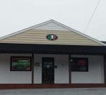 A small building with a beige exterior and a black awning. It has a sign featuring deli meat and cheese. There are two neon signs on the windows: one says "OPEN" and the other says "NADEAU'S SUBS AND DELI." The entrance door is centered between the windows.