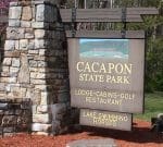 A wooden and stone sign at the entrance of Cacapon State Park. The sign lists amenities including lodge, cabins, golf, restaurant, lake, swimming, and horses. Trees and grass are visible in the background.