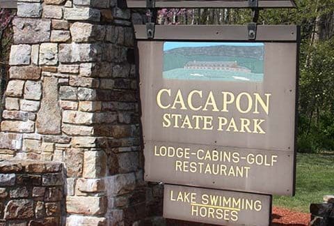A wooden and stone sign at the entrance of Cacapon State Park. The sign lists amenities including lodge, cabins, golf, restaurant, lake, swimming, and horses. Trees and grass are visible in the background.