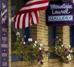 Exterior of a brick building with an American flag, window boxes displaying vibrant flowers, and signs reading "Fine American Crafts," "Jewelry," "Prints & Paintings," "Pottery," "Glass," and "Mountain Laurel Gallery.