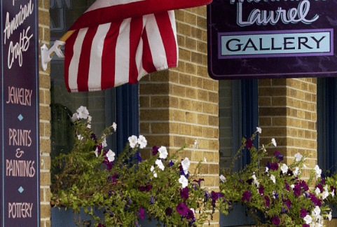 Exterior of a brick building with an American flag, window boxes displaying vibrant flowers, and signs reading "Fine American Crafts," "Jewelry," "Prints & Paintings," "Pottery," "Glass," and "Mountain Laurel Gallery.