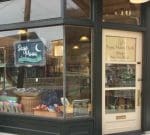 A storefront with a sign that reads "Sage Moon Herb Shop" above a crescent moon and star image. The shop window displays various items, including baskets and bags. The door, with the shop's name and hours written on it, is numbered 110.