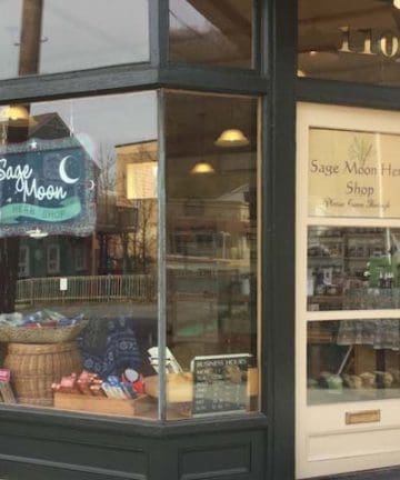 A storefront with a sign that reads "Sage Moon Herb Shop" above a crescent moon and star image. The shop window displays various items, including baskets and bags. The door, with the shop's name and hours written on it, is numbered 110.