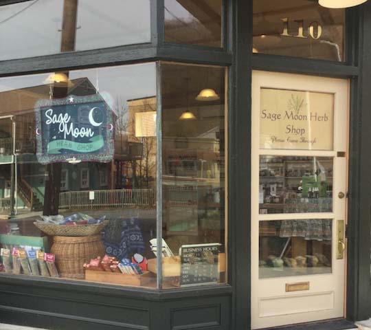 A storefront with a sign that reads "Sage Moon Herb Shop" above a crescent moon and star image. The shop window displays various items, including baskets and bags. The door, with the shop's name and hours written on it, is numbered 110.