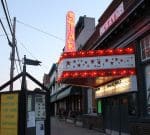 A street view of a vintage theater with a lit marquee reading "STAR." The theater's sign is adorned with red lights and stars. A signboard on the left advertises a free concert in the park every Saturday at 5:30 PM. Nearby buildings line the sidewalk.