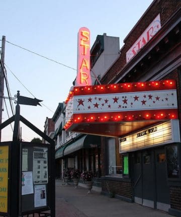 A street view of a vintage theater with a lit marquee reading "STAR." The theater's sign is adorned with red lights and stars. A signboard on the left advertises a free concert in the park every Saturday at 5:30 PM. Nearby buildings line the sidewalk.