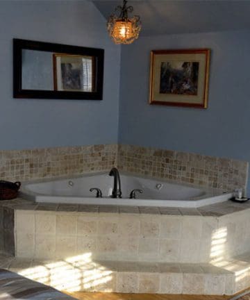 A tiled corner bathtub with a central faucet in a blue-walled room. A basket sits on the ledge beside the tub, and a small candle is on the other side. A mirror is mounted above the tub, and a framed painting hangs on the adjacent wall.