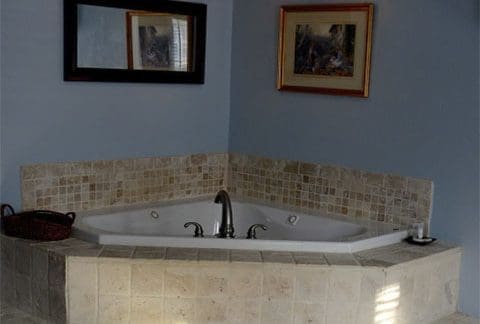 A tiled corner bathtub with a central faucet in a blue-walled room. A basket sits on the ledge beside the tub, and a small candle is on the other side. A mirror is mounted above the tub, and a framed painting hangs on the adjacent wall.