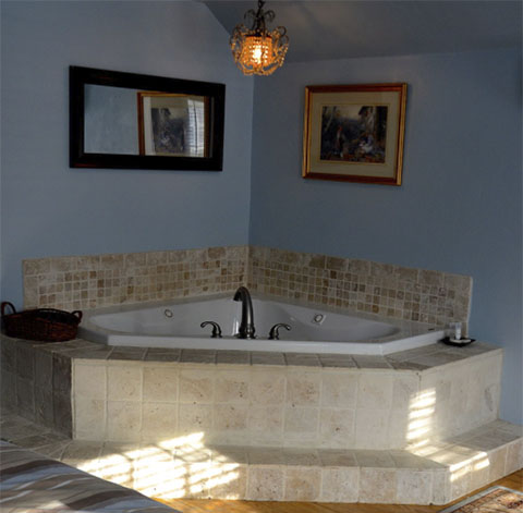 A tiled corner bathtub with a central faucet in a blue-walled room. A basket sits on the ledge beside the tub, and a small candle is on the other side. A mirror is mounted above the tub, and a framed painting hangs on the adjacent wall.