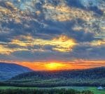 A vibrant sunset illuminates the sky with warm hues of orange and yellow over a lush, green, hilly landscape. The scene includes a white farmhouse nestled among trees and rolling hills, with a dramatic, cloud-filled sky creating a striking backdrop.