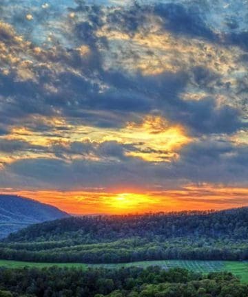 A vibrant sunset illuminates the sky with warm hues of orange and yellow over a lush, green, hilly landscape. The scene includes a white farmhouse nestled among trees and rolling hills, with a dramatic, cloud-filled sky creating a striking backdrop.