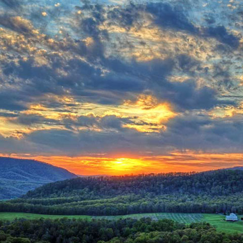 A vibrant sunset illuminates the sky with warm hues of orange and yellow over a lush, green, hilly landscape. The scene includes a white farmhouse nestled among trees and rolling hills, with a dramatic, cloud-filled sky creating a striking backdrop.