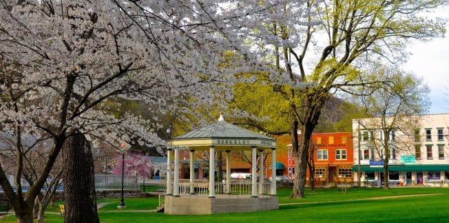 A picturesque park scene featuring a small white gazebo labeled "Berkeley Springs." Blooming cherry blossom trees frame the area, with colorful historic buildings in the background. The lush green grass and large trees add to the serene atmosphere.