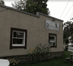 A beige building with a sign that reads "Ravenwood." A window sign says "Yes We’re Open." There are sunflowers growing near the building and a picnic table in front. A road and houses are visible in the background.