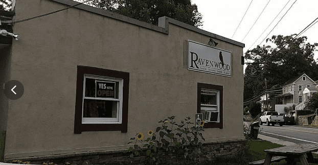 A beige building with a sign that reads "Ravenwood." A window sign says "Yes We’re Open." There are sunflowers growing near the building and a picnic table in front. A road and houses are visible in the background.
