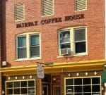 A two-story brick building houses Fairfax Coffee House. The entrance features yellow trim and large windows with a small, square canopy. "Fairfax Coffee House" is written in large letters above the second-story windows. Yellow outdoor chairs are in front.