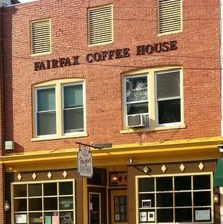 A two-story brick building houses Fairfax Coffee House. The entrance features yellow trim and large windows with a small, square canopy. "Fairfax Coffee House" is written in large letters above the second-story windows. Yellow outdoor chairs are in front.