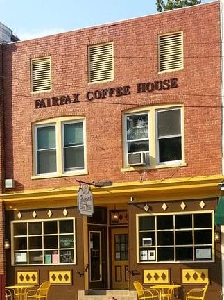 A two-story brick building houses Fairfax Coffee House. The entrance features yellow trim and large windows with a small, square canopy. "Fairfax Coffee House" is written in large letters above the second-story windows. Yellow outdoor chairs are in front.