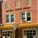 A two-story brick building houses Fairfax Coffee House. The entrance features yellow trim and large windows with a small, square canopy. "Fairfax Coffee House" is written in large letters above the second-story windows. Yellow outdoor chairs are in front.