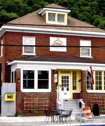 A two-story red brick building with a tan roof, white-framed windows, and a small porch. It has a sign above the porch with a logo and an American flag by the entrance. There are outdoor chairs and tables in front, along with parking spaces and greenery in the background.