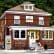 A two-story red brick building with a tan roof, white-framed windows, and a small porch. It has a sign above the porch with a logo and an American flag by the entrance. There are outdoor chairs and tables in front, along with parking spaces and greenery in the background.