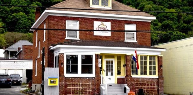 A two-story red brick building with a tan roof, white-framed windows, and a small porch. It has a sign above the porch with a logo and an American flag by the entrance. There are outdoor chairs and tables in front, along with parking spaces and greenery in the background.