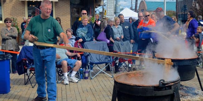 A man wearing a green shirt and blue jeans stirs a large black pot with a wooden paddle at an outdoor event. A crowd of people watches, seated and standing around the area. The event takes place near a brick building with a sign "Mountain" and steam rises from the pots.