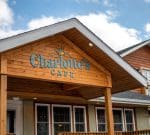 A building features a sign with the name "Charlotte's Cafe" on a wooden peaked roof. The facade has large windows and a covered porch. The sky is partly cloudy, suggesting a bright day.