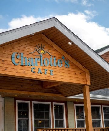 A building features a sign with the name "Charlotte's Cafe" on a wooden peaked roof. The facade has large windows and a covered porch. The sky is partly cloudy, suggesting a bright day.