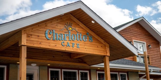 A building features a sign with the name "Charlotte's Cafe" on a wooden peaked roof. The facade has large windows and a covered porch. The sky is partly cloudy, suggesting a bright day.