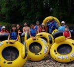 A group of people in life vests stand and sit on large yellow inflatable tubes labeled "RAPID RIDER" by a riverbank with a lush green forest in the background. Everyone appears ready for a river tubing adventure.