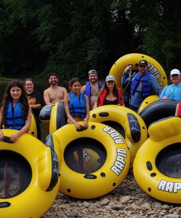 A group of people in life vests stand and sit on large yellow inflatable tubes labeled "RAPID RIDER" by a riverbank with a lush green forest in the background. Everyone appears ready for a river tubing adventure.