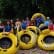 A group of people in life vests stand and sit on large yellow inflatable tubes labeled "RAPID RIDER" by a riverbank with a lush green forest in the background. Everyone appears ready for a river tubing adventure.