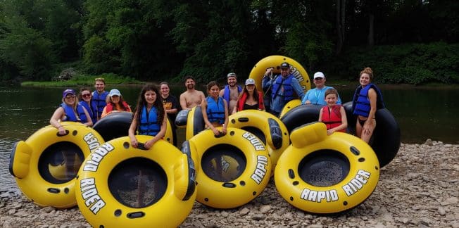 A group of people in life vests stand and sit on large yellow inflatable tubes labeled "RAPID RIDER" by a riverbank with a lush green forest in the background. Everyone appears ready for a river tubing adventure.
