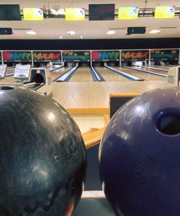 Two bowling balls, one black and one purple, rest on a ball return rack. In the background, several bowling lanes extend into the distance, with bright digital scoring screens overhead. The setting appears to be a well-lit and colorful bowling alley.