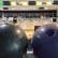 Two bowling balls, one black and one purple, rest on a ball return rack. In the background, several bowling lanes extend into the distance, with bright digital scoring screens overhead. The setting appears to be a well-lit and colorful bowling alley.