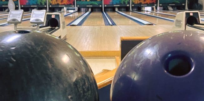 Two bowling balls, one black and one purple, rest on a ball return rack. In the background, several bowling lanes extend into the distance, with bright digital scoring screens overhead. The setting appears to be a well-lit and colorful bowling alley.
