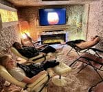 Three people relax in recliner chairs in a dimly lit room, simulating a cave-like environment with textured walls. They are watching a screen displaying a warm, glowing image. A modern electric fireplace adds to the cozy ambiance.
