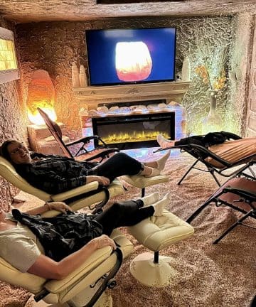 Three people relax in recliner chairs in a dimly lit room, simulating a cave-like environment with textured walls. They are watching a screen displaying a warm, glowing image. A modern electric fireplace adds to the cozy ambiance.
