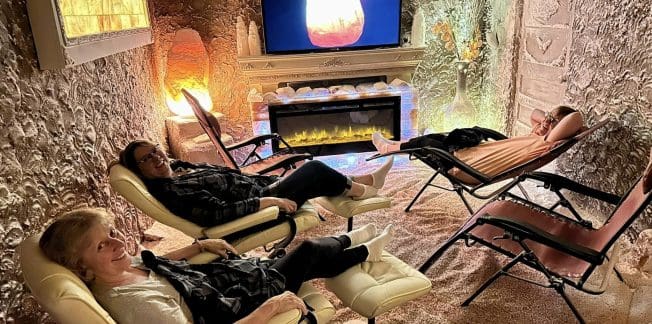 Three people relax in recliner chairs in a dimly lit room, simulating a cave-like environment with textured walls. They are watching a screen displaying a warm, glowing image. A modern electric fireplace adds to the cozy ambiance.