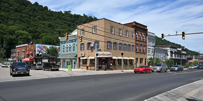 A small town street scene with a row of historic buildings, including a brick corner building with a restaurant. Several cars and a bicycle are on the street. There are trees and hills in the background under a blue sky with some clouds.