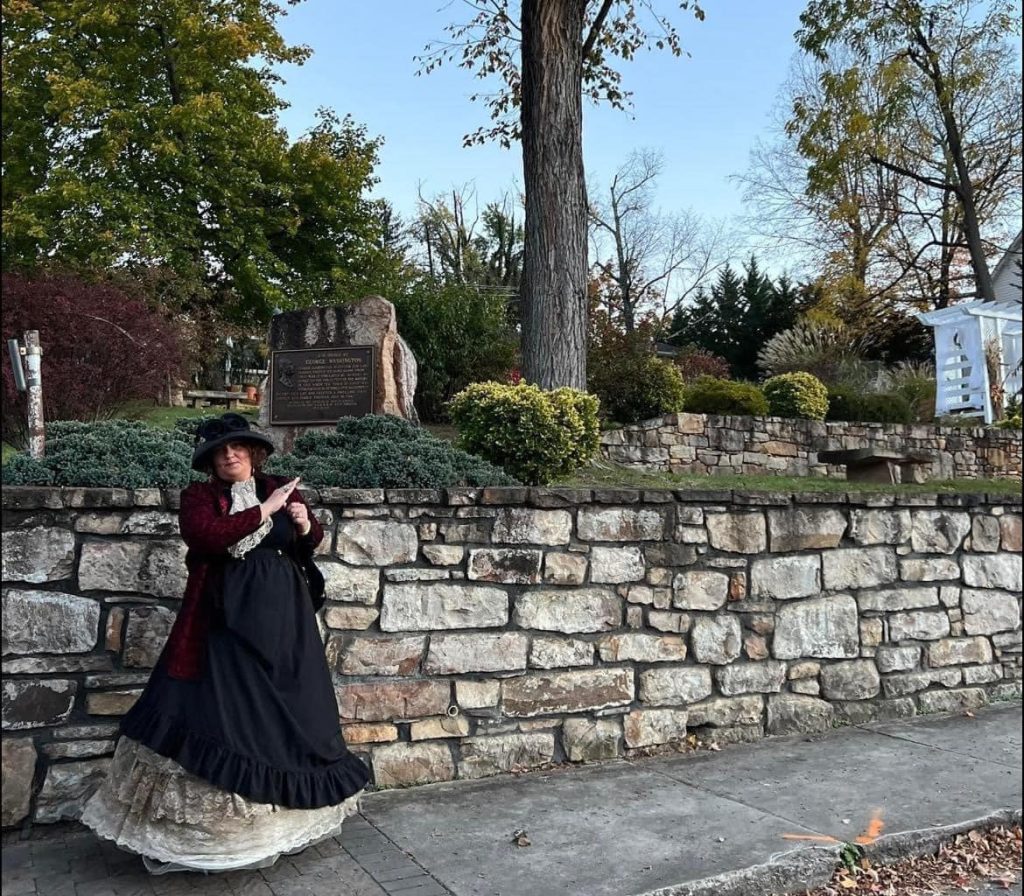 A person dressed in historical attire, including a black hat and a layered dress, stands by a stone wall in a park. There are trees and a plaque in the background, and the setting appears to be during autumn with fallen leaves on the ground.