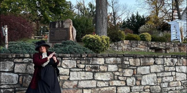 A person dressed in historical attire, including a black hat and a layered dress, stands by a stone wall in a park. There are trees and a plaque in the background, and the setting appears to be during autumn with fallen leaves on the ground.