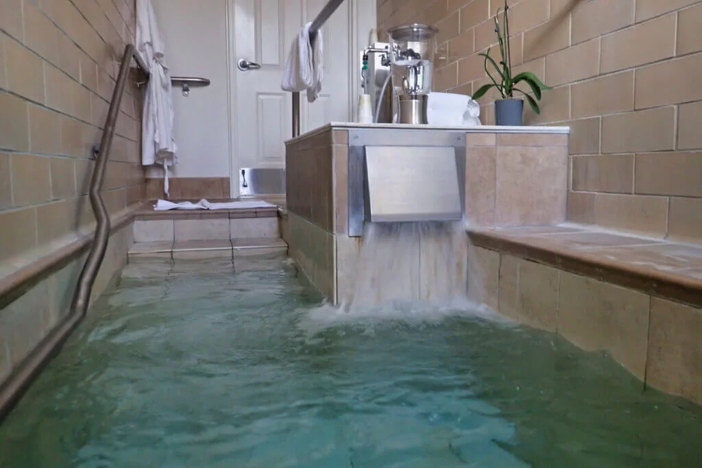 A tranquil indoor spa room with a small pool. Water pours from a stainless steel spout into the pool, surrounded by beige tiles. A towel hangs on the wall, and a potted plant adds a touch of greenery to the scene.