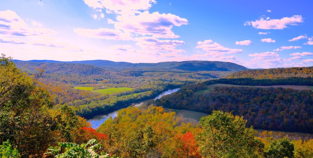 A scenic view of a winding river surrounded by vibrant autumn trees and rolling hills under a blue sky with scattered clouds. The landscape features lush greenery mixed with red, orange, and yellow foliage.